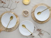 Draufsicht auf ein minimalistisches Tisch-Arrangement mit drei weißen Keramikschalen auf runden Holzbrettchen, jeweils mit goldfarbenem Löffel, auf hellem Leinentuch mit Beerenzweigen und einem Glas mit gelber Flüssigkeit.