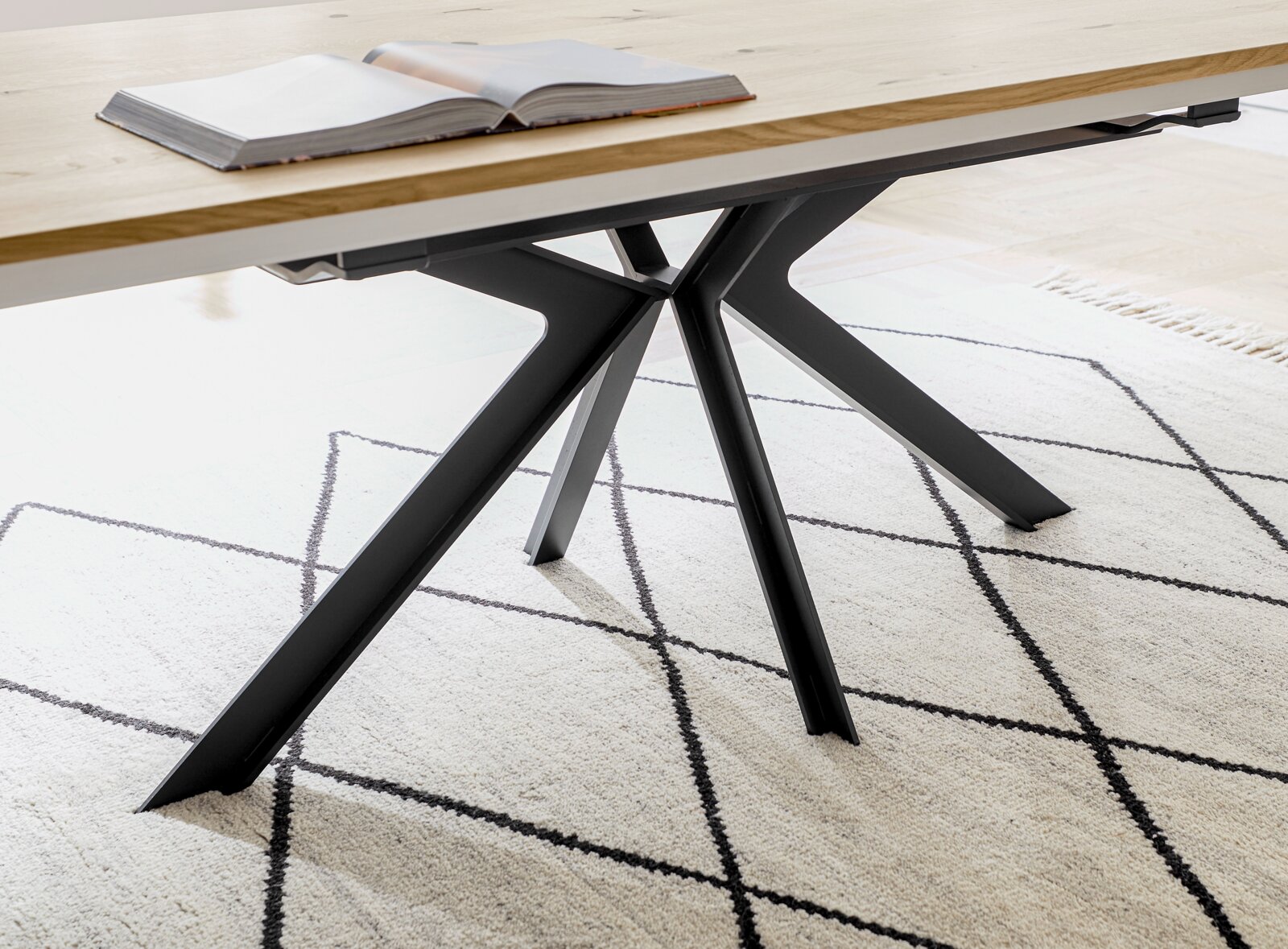 Detailaufnahme in schräger Seitenperspektive: moderner Esstisch mit heller Holzplatte und schwarzem Metallgestell in Sternform auf gemustertem Teppich, mit aufgeschlagenem Buch auf der Tischplatte