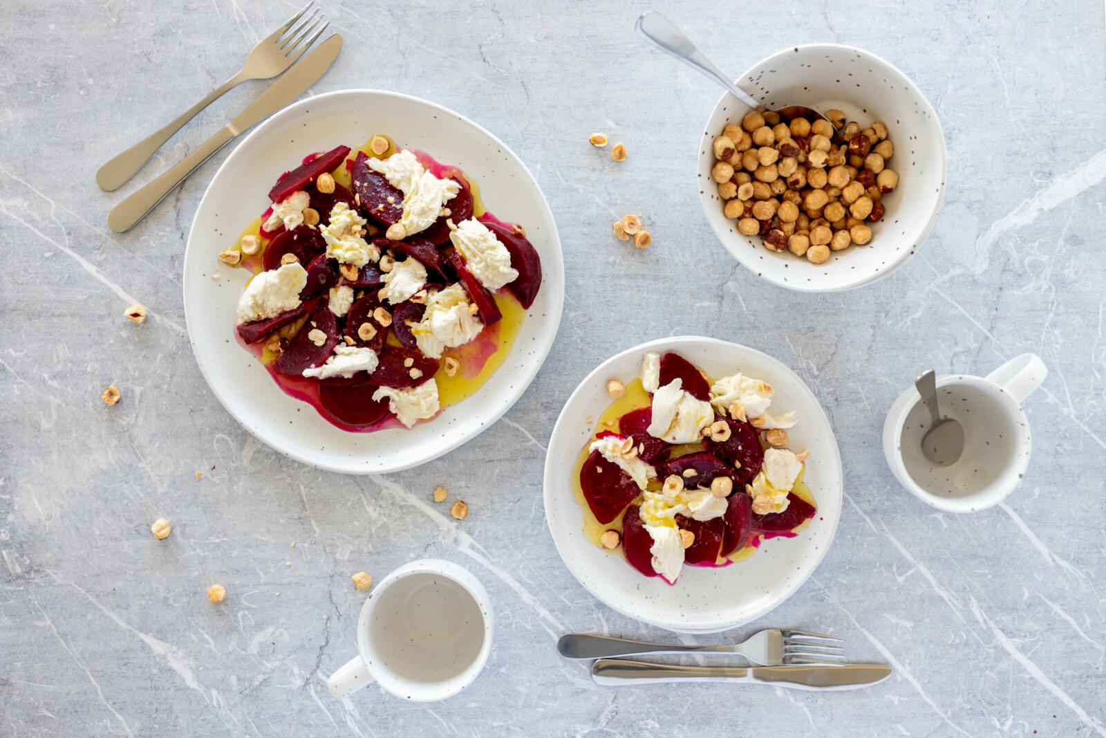 Draufsicht auf zwei Teller mit Rote-Bete-Salat, cremigem Käse, Haselnüssen und Olivenöl auf hellem Steintisch, daneben Schale mit Haselnüssen und Besteck