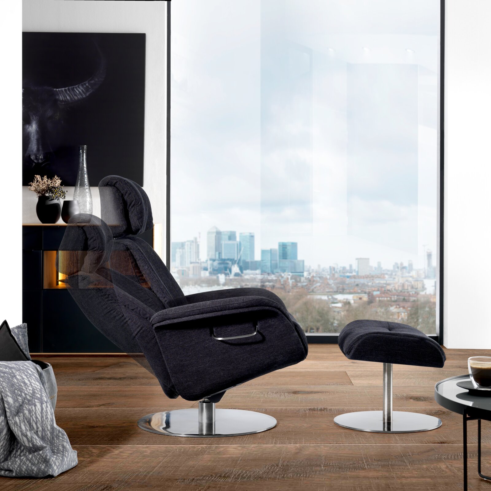 Moderne relaxfauteuil met voetenbankje, bekleed met donkere stof, gezien vanaf de zijkant, voor een groot raam met uitzicht op de stad.