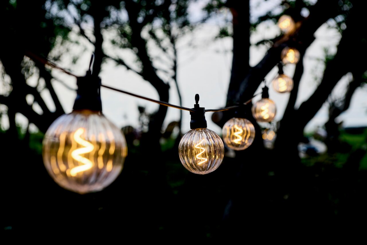 Sirius LED-lichtslinger voor buiten, 10 m, 10 LED's, verlengstuk LUKE Nahaufnahme einer Lichterkette mit klaren, runden LED-Glühbirnen, aufgehängt zwischen Bäumen in einem Garten bei Dämmerung.