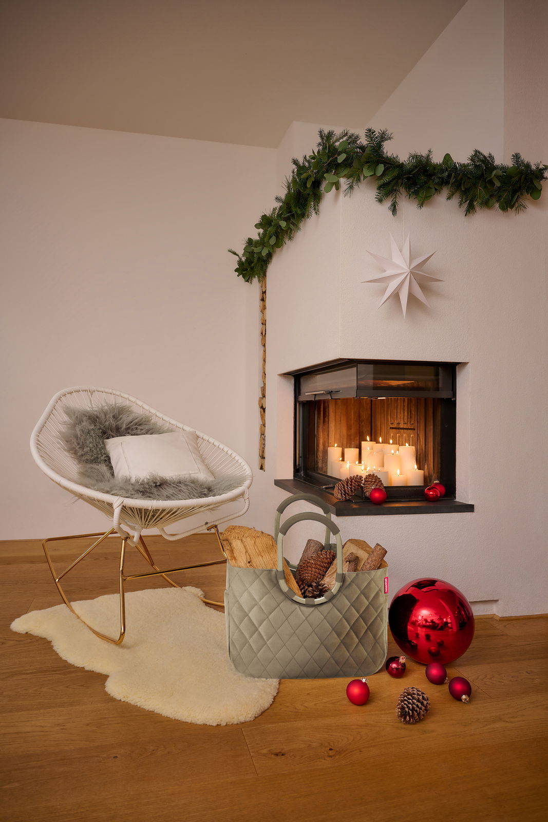 Ein stilvoller, grauer loopshopper L Korb steht auf einem Holzboden vor einem gemütlichen Kamin mit brennenden Kerzen. Der Korb ist mit Holzscheiten und Tannenzapfen gefüllt. Im Hintergrund ist ein moderner, weißer Schaukelstuhl mit einem Kissen und einem