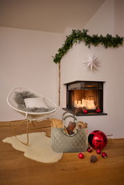 Ein stilvoller, grauer loopshopper L Korb steht auf einem Holzboden vor einem gemütlichen Kamin mit brennenden Kerzen. Der Korb ist mit Holzscheiten und Tannenzapfen gefüllt. Im Hintergrund ist ein moderner, weißer Schaukelstuhl mit einem Kissen und einem