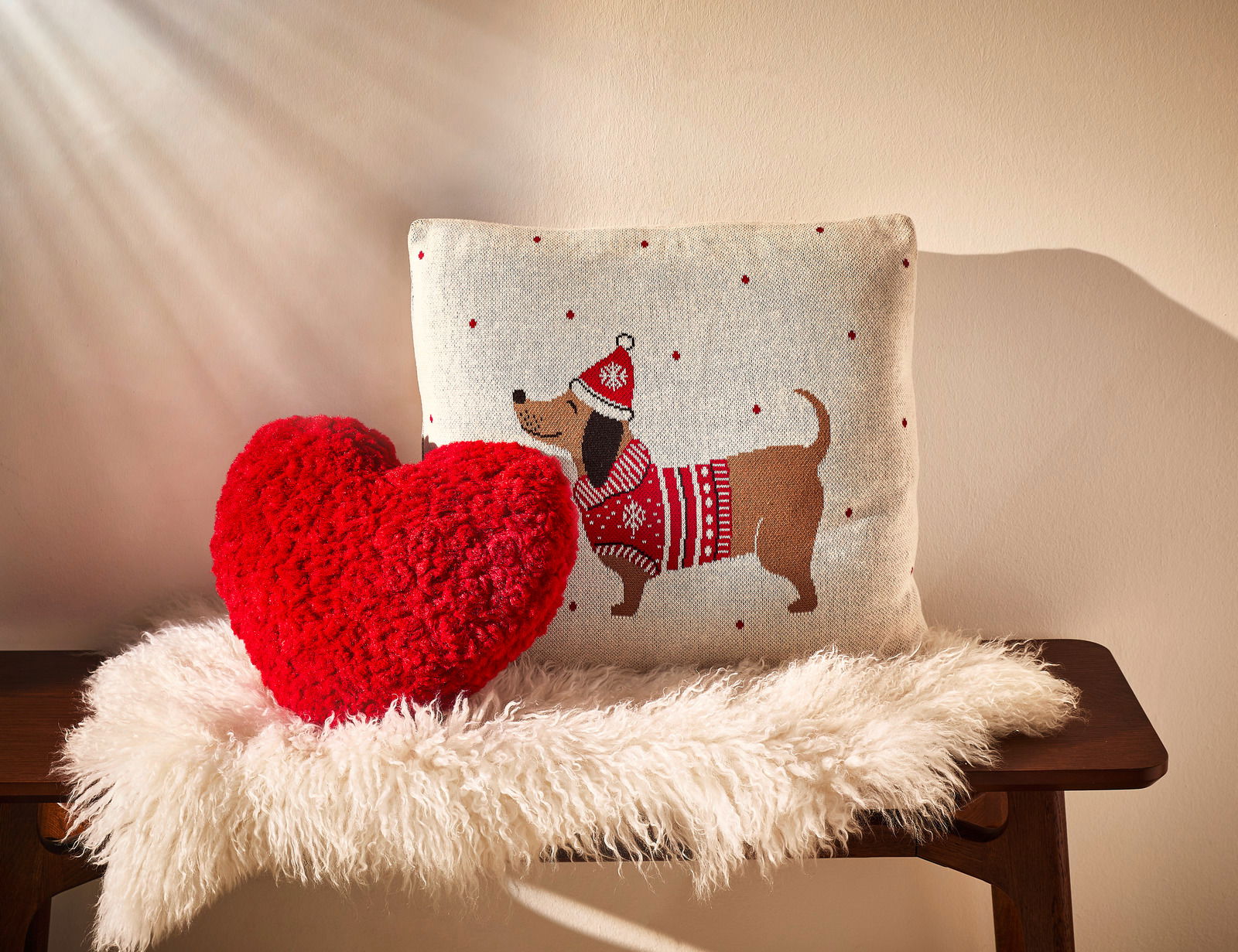 Zwei Zierkissen auf einer Bank, eines in Herzform aus rotem Plüsch und das andere mit einem Hundemotiv in Winterkleidung, von vorne fotografiert.