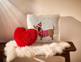 Zwei Zierkissen auf einer Bank, eines in Herzform aus rotem Plüsch und das andere mit einem Hundemotiv in Winterkleidung, von vorne fotografiert.