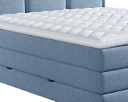 Close-up vanuit schuin vooraanzicht van een blauw gestoffeerd boxspringbed met een dikke, wit doorgestikte matrastopper.