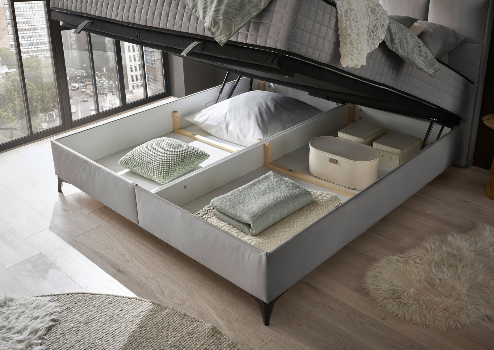 Boxbett mit geöffnetem Stauraum, gefüllt mit Kissen und Decken, aus der Perspektive eines Fensters mit Stadtblick.