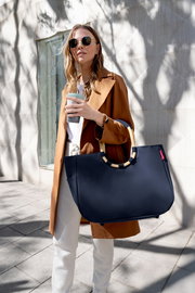 Frau in stilvoller Kleidung trägt eine große, dunkelblaue Tasche mit runden Griffen, während sie einen Kaffee hält. Die Tasche wird von der Seite gezeigt.