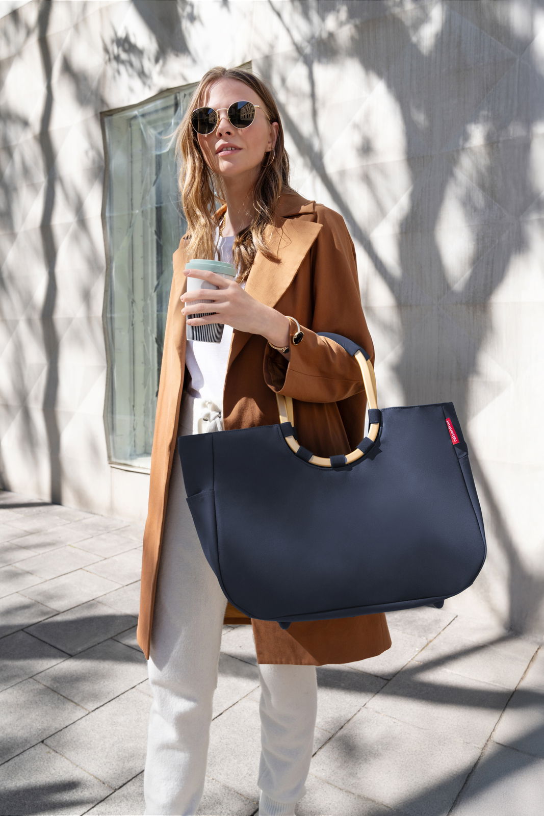 Frau in stilvoller Kleidung trägt eine große, dunkelblaue Tasche mit runden Griffen, während sie einen Kaffee hält. Die Tasche wird von der Seite gezeigt.