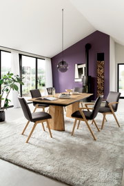 Moderne eethoek met rechthoekige houten eettafel en vier grijze stoelen op licht vloerkleed, met glazen hanglamp en paarse accentmuur op de achtergrond, gefotografeerd in schuin vooraanzicht