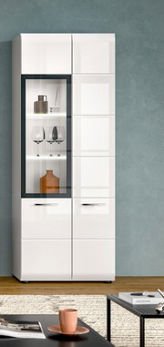 Moderne weiße Hochglanz-Vitrine/Schrank mit linksseitiger Glastür und beleuchteten Glasböden, darin Gläser und Deko, vor grauer Wand; frontale Ansicht.