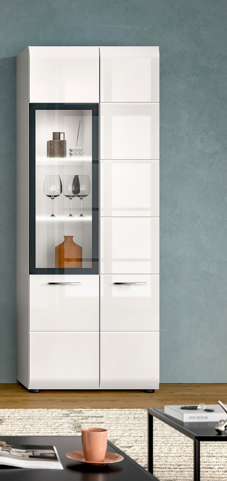 Moderne weiße Hochglanz-Vitrine/Schrank mit linksseitiger Glastür und beleuchteten Glasböden, darin Gläser und Deko, vor grauer Wand; frontale Ansicht.