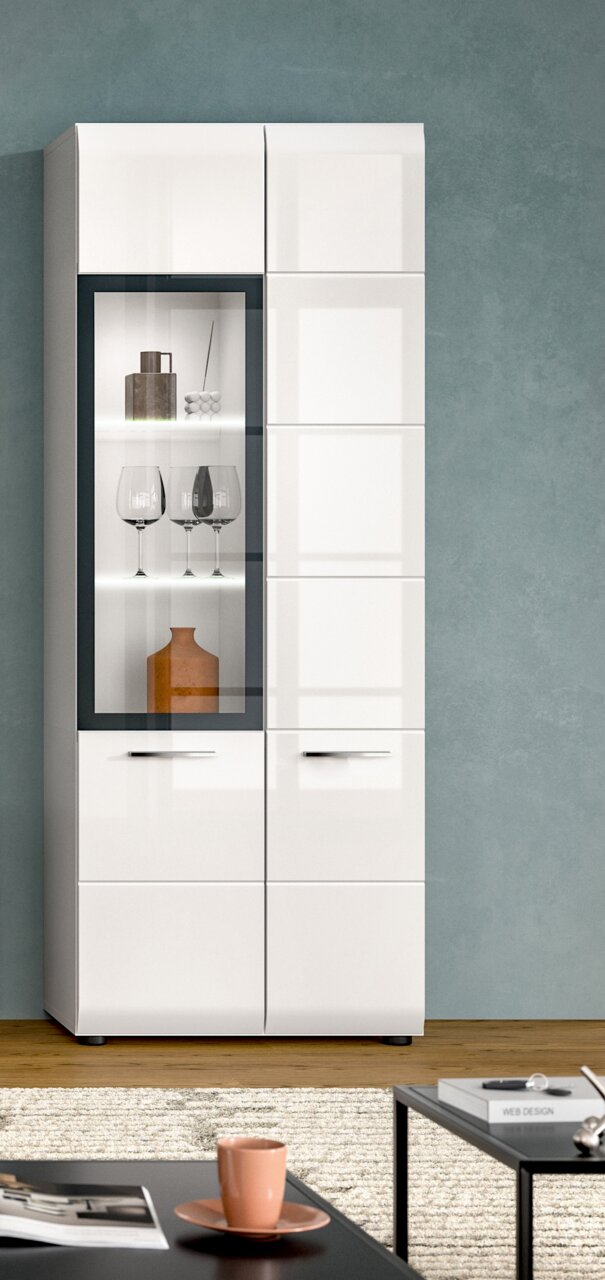 Moderne weiße Hochglanz-Vitrine/Schrank mit linksseitiger Glastür und beleuchteten Glasböden, darin Gläser und Deko, vor grauer Wand; frontale Ansicht.