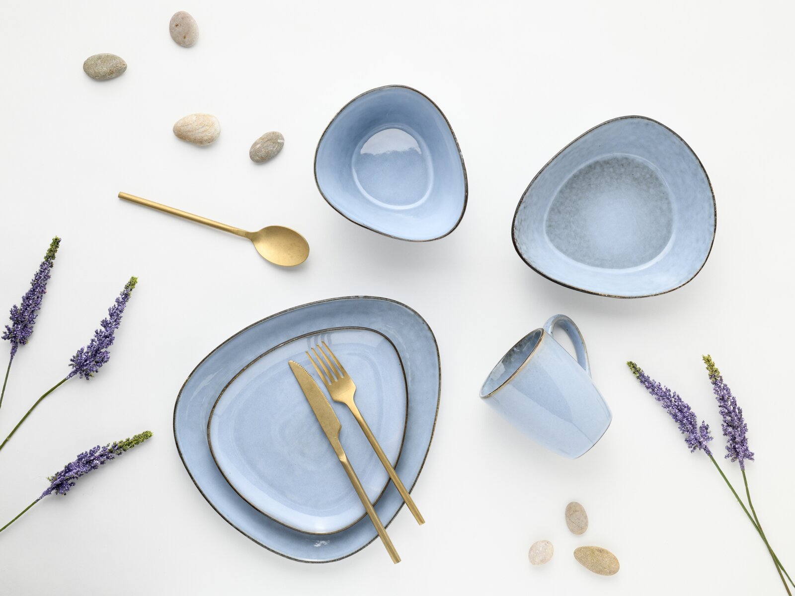 Hellblaues Keramik-Geschirrset mit Teller, Schalen und Tasse, kombiniert mit goldfarbenem Besteck auf weißem Hintergrund, Draufsicht von oben (Flatlay) mit Lavendelzweigen und dekorativen Steinen.