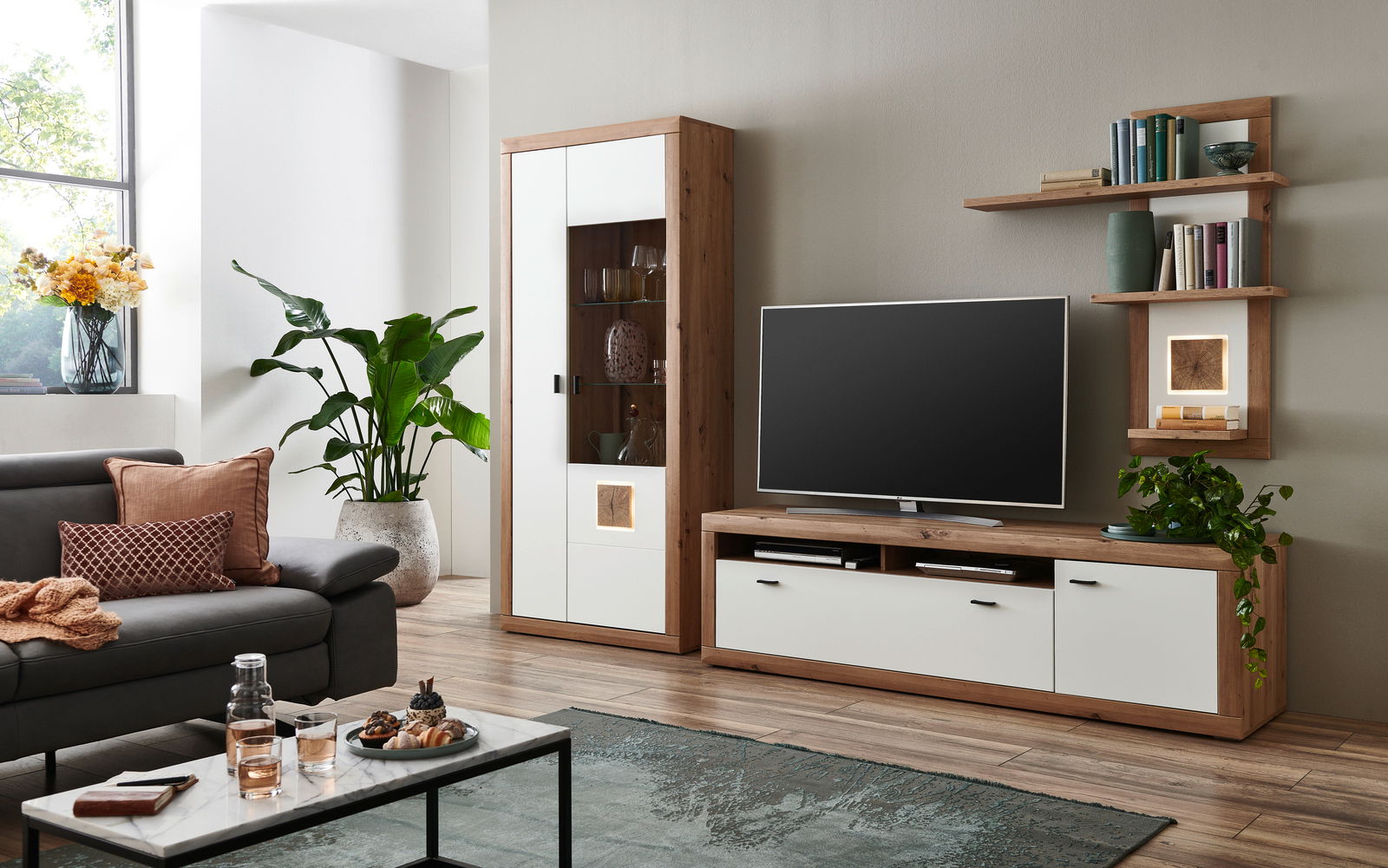 Moderne Wohnzimmer-Wandkombination in Holzoptik mit weißem Akzent, bestehend aus einem TV-Schrank, einem hohen Schrank mit Glastür und Regalen, aus der Perspektive des Wohnzimmers betrachtet.