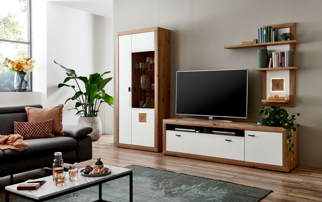 Moderne woonkamerwandcombinatie in houtlook met witte accenten, bestaande uit een tv-kast, een hoge kast met glazen deur en planken, gezien vanuit de woonkamer.