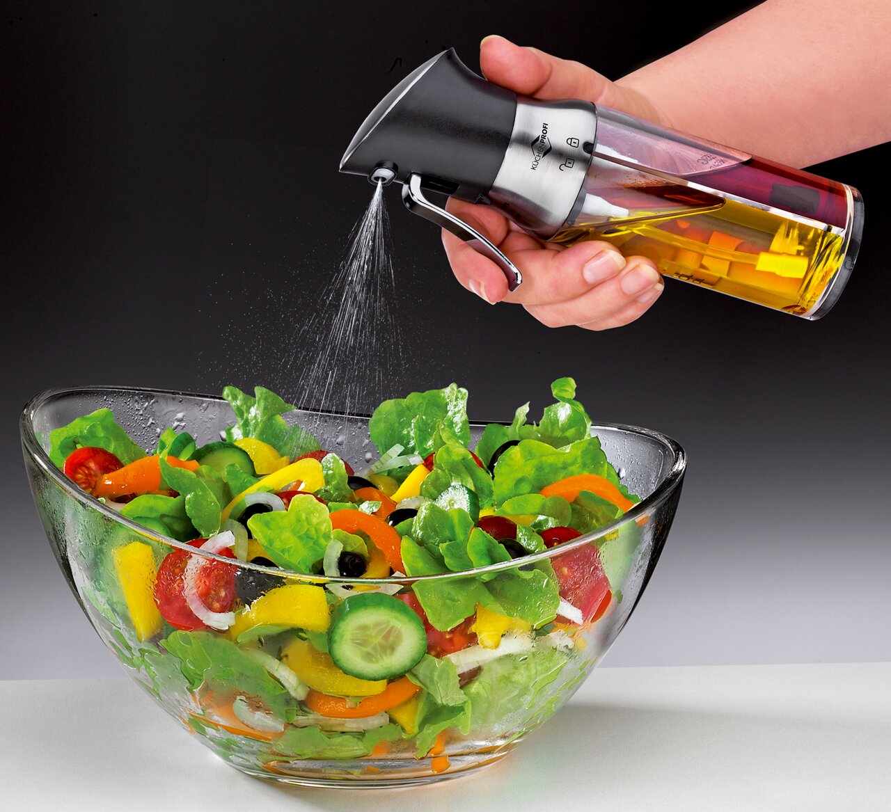 Een azijnspuit/oliedispenser in de hand, waarmee olie over een verse salade in een glazen schaal wordt gespoten, gezien vanuit de zijkant.