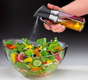 Een azijnspuit/oliedispenser in de hand, waarmee olie over een verse salade in een glazen schaal wordt gespoten, gezien vanuit de zijkant.