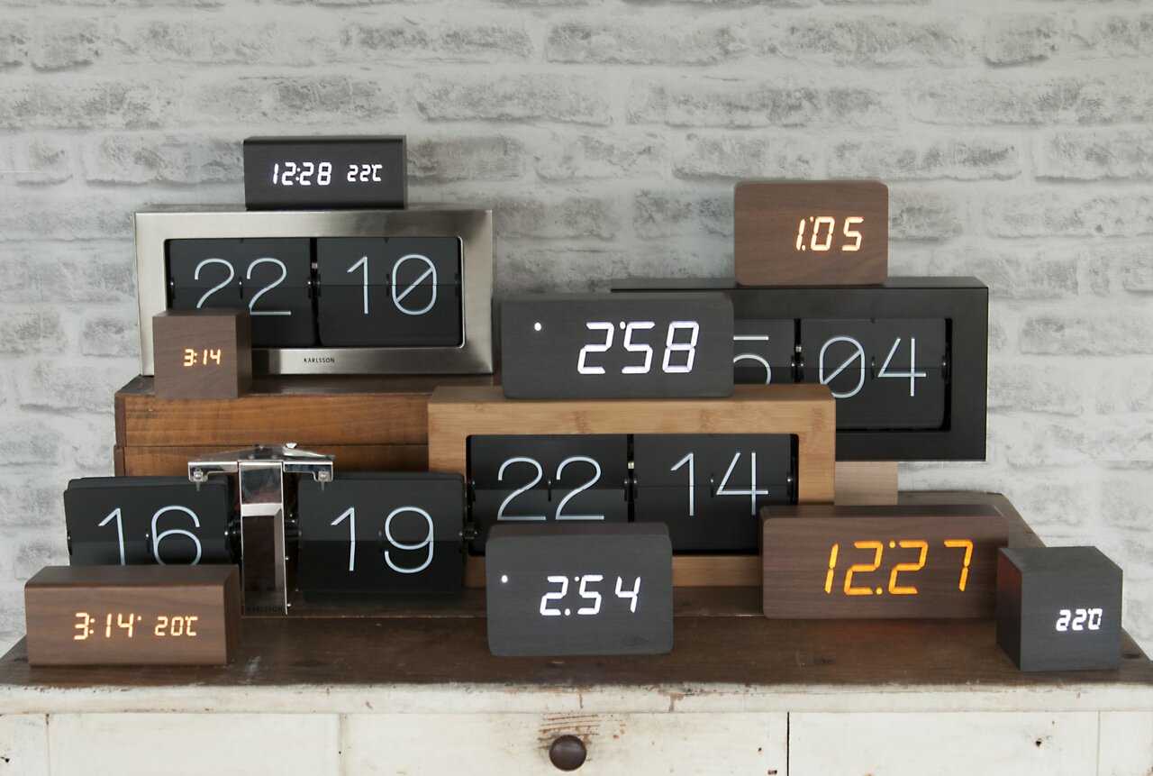Verschillende moderne klokken van hout en metaal, gezien van voren, die op een tafel voor een bakstenen muur staan.