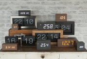 Verschiedene moderne Flip-Uhren aus Holz und Metall in einer frontalen Ansicht, die auf einem Tisch vor einer Ziegelwand angeordnet sind.