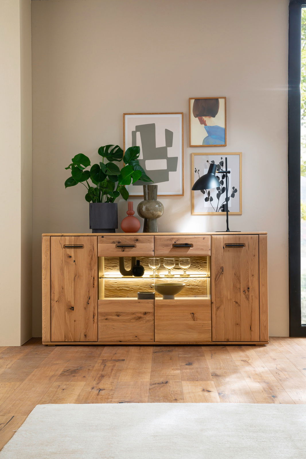 Holz-Sideboard mit Glasvitrine, dekoriert mit Pflanzen und Kunstwerken, aus frontaler Perspektive.