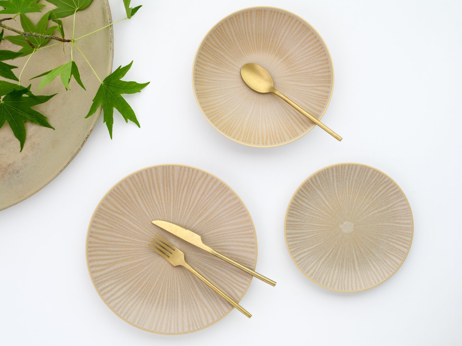 Beige Keramikteller-Set mit feiner radialer Rillenstruktur, arrangiert mit goldfarbenem Besteck auf weißem Hintergrund, Draufsicht von oben.