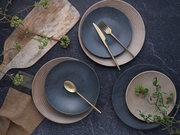 Vogelperspektive auf ein stilvolles 18-teiliges Tellerset mit blauen und beigen Tellern, dekoriert mit goldfarbenem Besteck und grünen Zweigen auf einer dunklen Steinoberfläche.