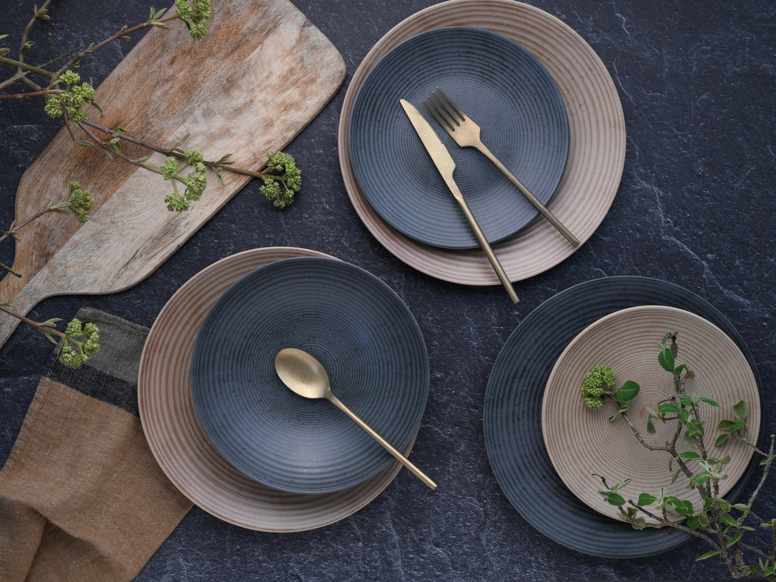 Vogelperspektive auf ein stilvolles 18-teiliges Tellerset mit blauen und beigen Tellern, dekoriert mit goldfarbenem Besteck und grünen Zweigen auf einer dunklen Steinoberfläche.