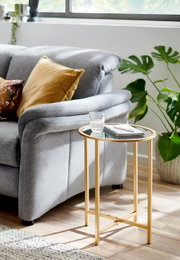 Runder Beistelltisch mit goldfarbenem Metallgestell und Glasplatte neben grauem Sofa, darauf Glas und Notizbuch; Perspektive: seitliche 3/4-Ansicht leicht von oben