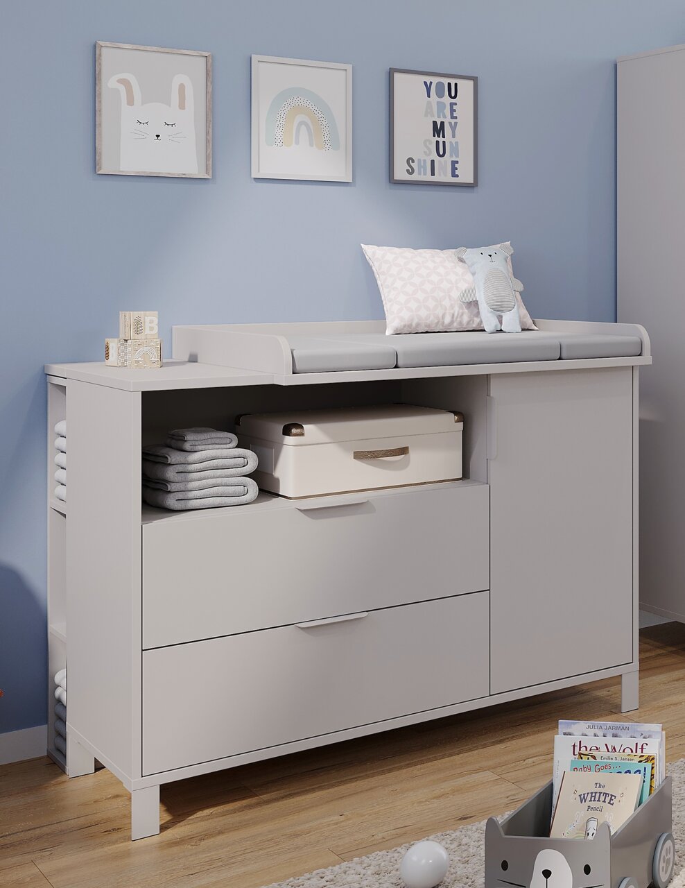 Weiße Wickelkommode in einem Babyzimmer, frontal fotografiert, mit Schubladen, Regalfächern und Dekorationen wie einem Kuscheltier und gerollten Handtüchern.