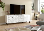 Weißes TV-Lowboard mit geometrisch strukturierter Front, Schubladen und Tür auf schwarzen Metallfüßen, mit Fernseher darauf, in moderner Wohnzimmerszene aus frontaler leicht seitlicher Perspektive.