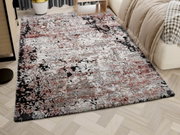 Designer Teppich in einem modernen Schlafzimmer, mit abstraktem Muster in Rot-, Grau- und Schwarztönen, auf hellem Holzboden neben einem Bett. Perspektive von schräg oben.