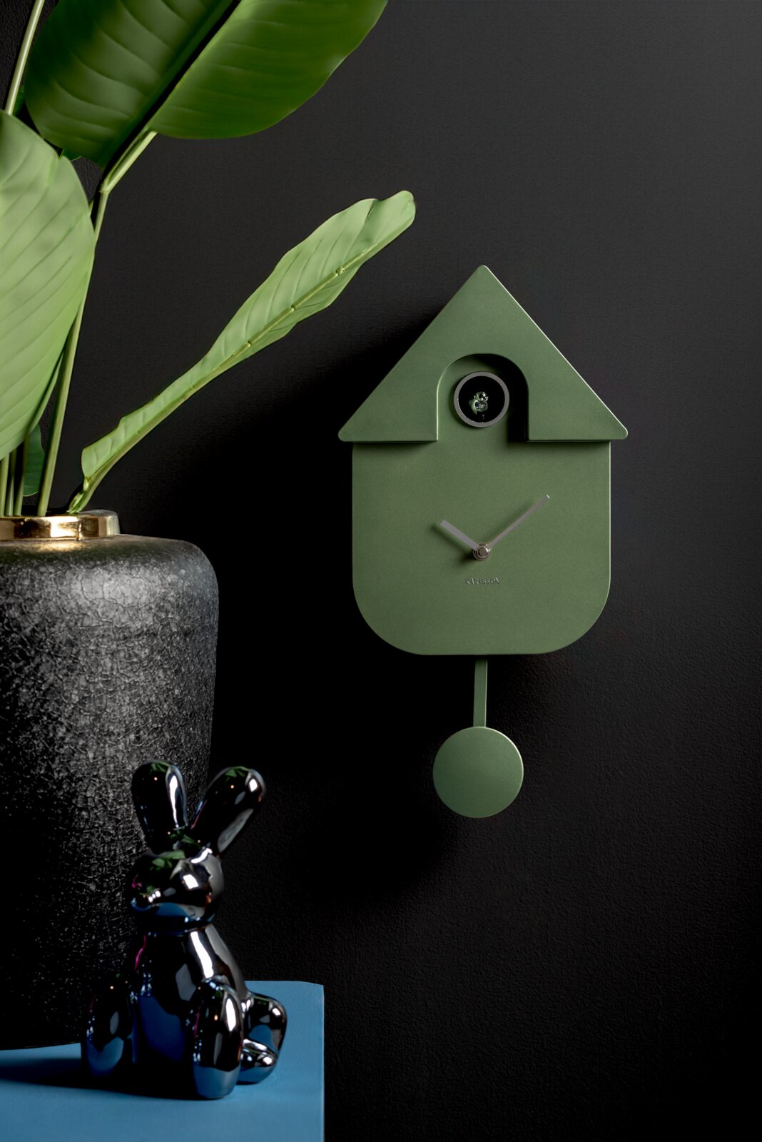 KARLSSON Wandklok CUCKOO Groene wandklok in de vorm van een vogelhuisje met slinger, gezien vanuit de zijkant, naast een plant en een zwart konijntje.
