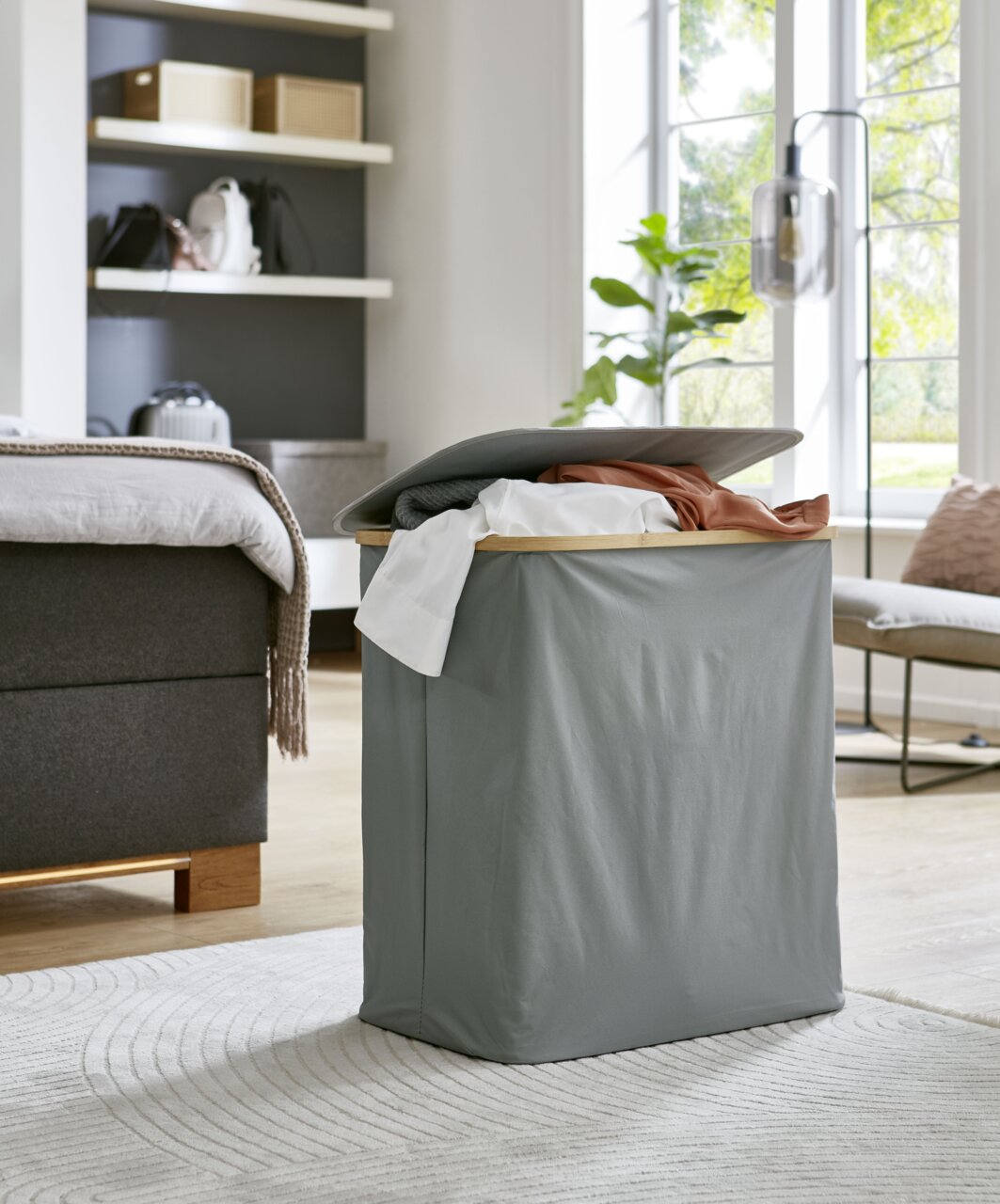 Grauer Wäschekorb mit Deckel und Holzrahmen, gefüllt mit Kleidung, im Schlafzimmer auf Teppich – Frontansicht in leicht schräger Perspektive.