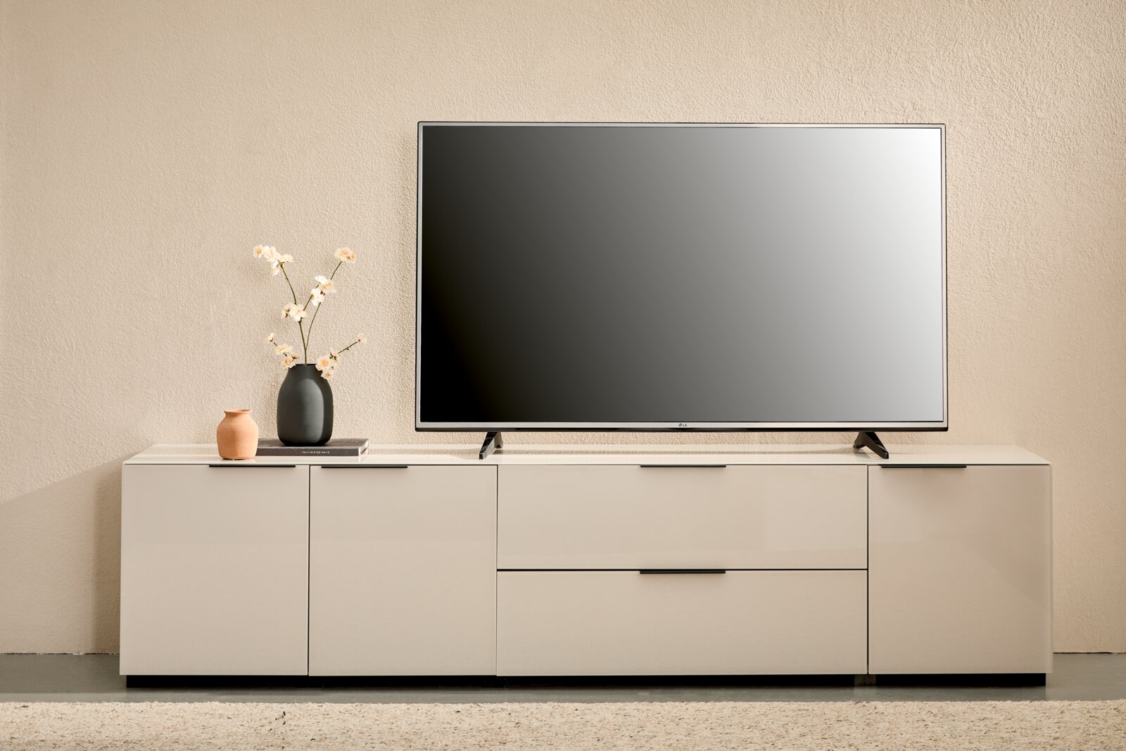 Novel XL-TV-Lowboard MALIN 2.0 Modernes weißes TV-Lowboard mit Flachbildfernseher, Vase mit Blüten und kleiner Deko-Vase auf minimalistischer Wohnzimmereinrichtung, Frontalansicht.