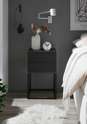 Moderne schwarze Nachtkommode mit geriffelter Oberfläche, dekoriert mit einer Vase und einer Uhr, seitliche Perspektive.