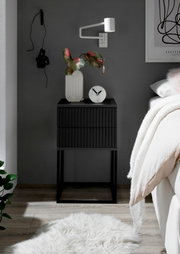 Moderne schwarze Nachtkommode mit geriffelter Oberfläche, dekoriert mit einer Vase und einer Uhr, seitliche Perspektive.