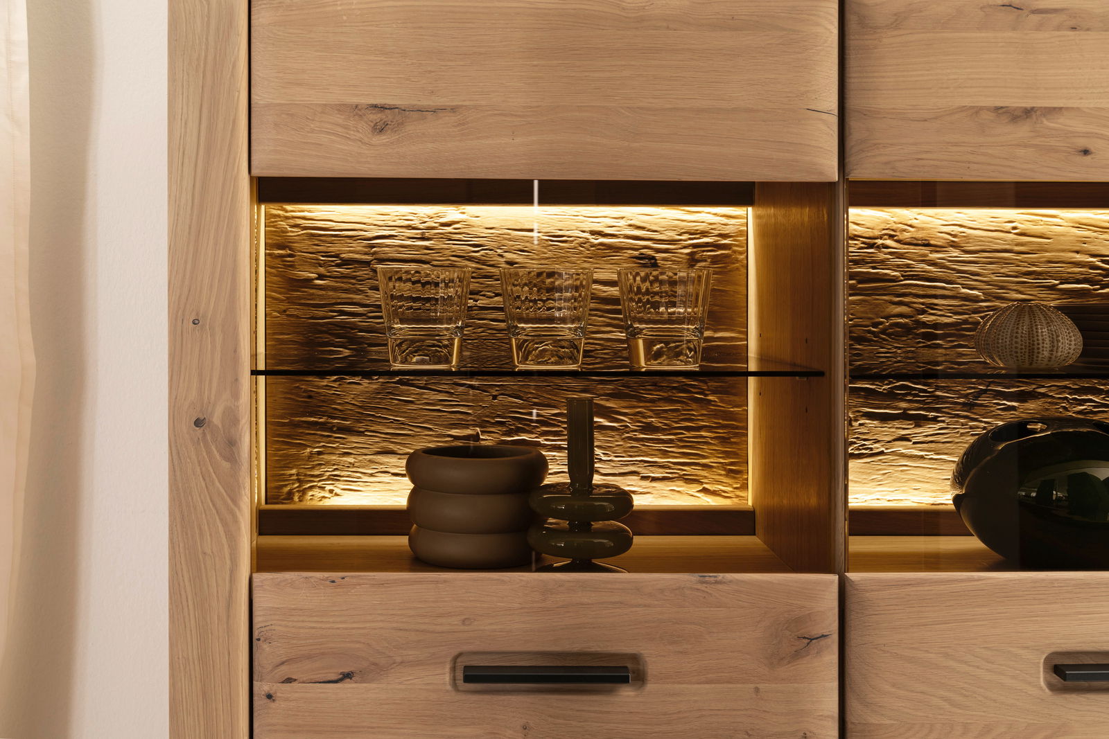 Nahaufnahme einer beleuchteten Wandkombination aus Holz mit Glasregal, auf dem Gläser und dekorative Objekte stehen.
