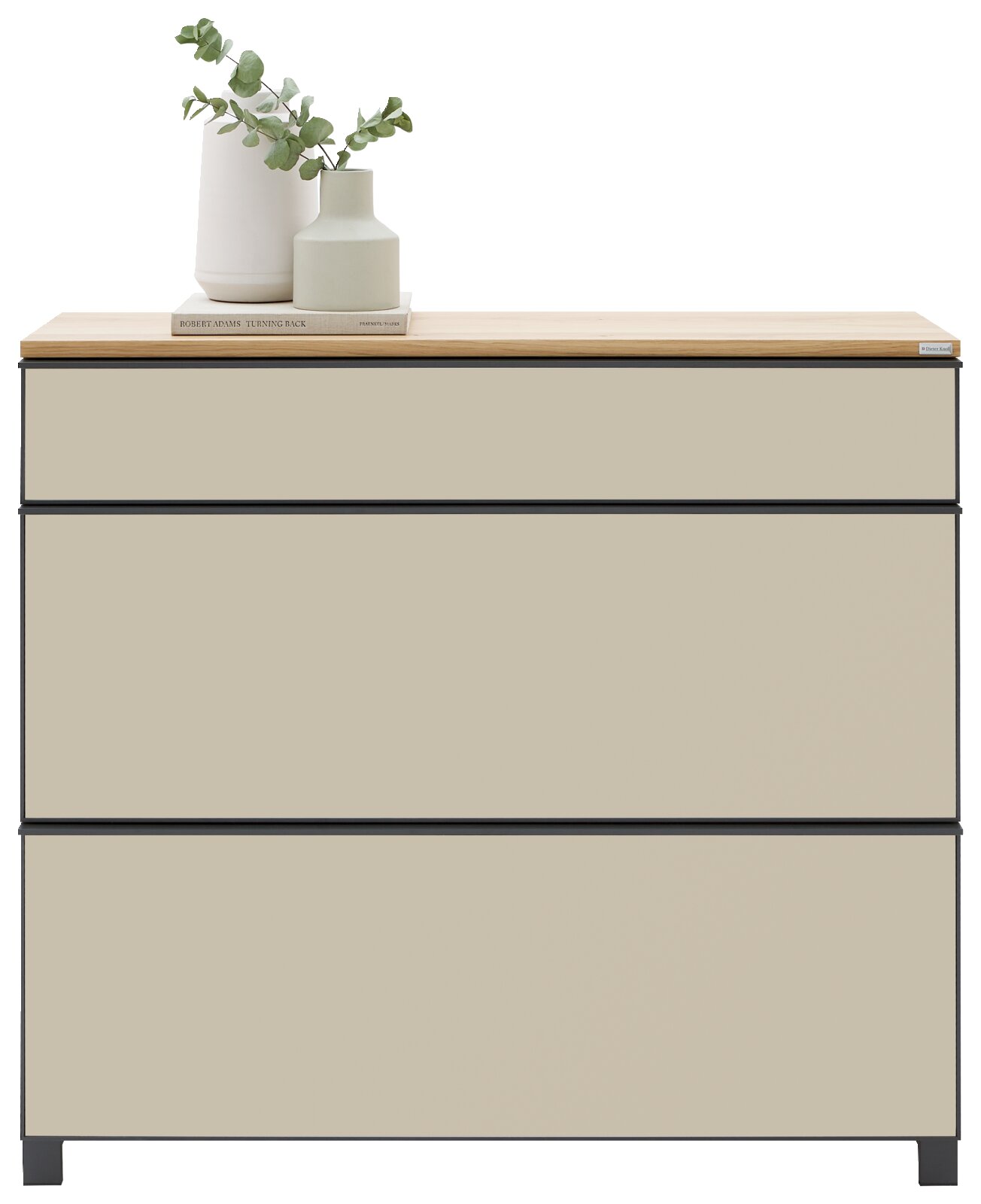 Moderne Kommode mit drei breiten Schubladen, beigem Korpus, schwarzem Rahmen und heller Holzplatte; dekoriert mit Vase, Buch und Eukalyptuszweig, Frontansicht auf weißem Hintergrund