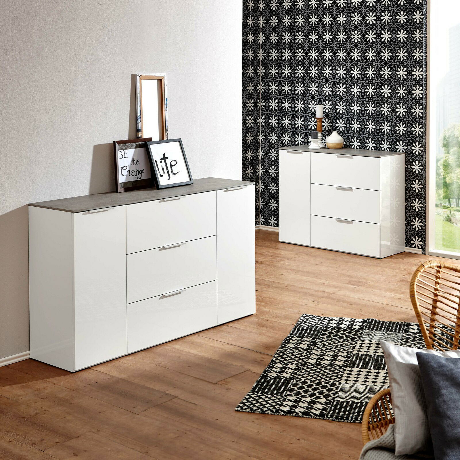 Modernes weißes Sideboard mit drei Schubladen und zwei Türen, in einem stilvoll eingerichteten Raum mit Holzboden und gemustertem Teppich, aus einer seitlichen Perspektive fotografiert.