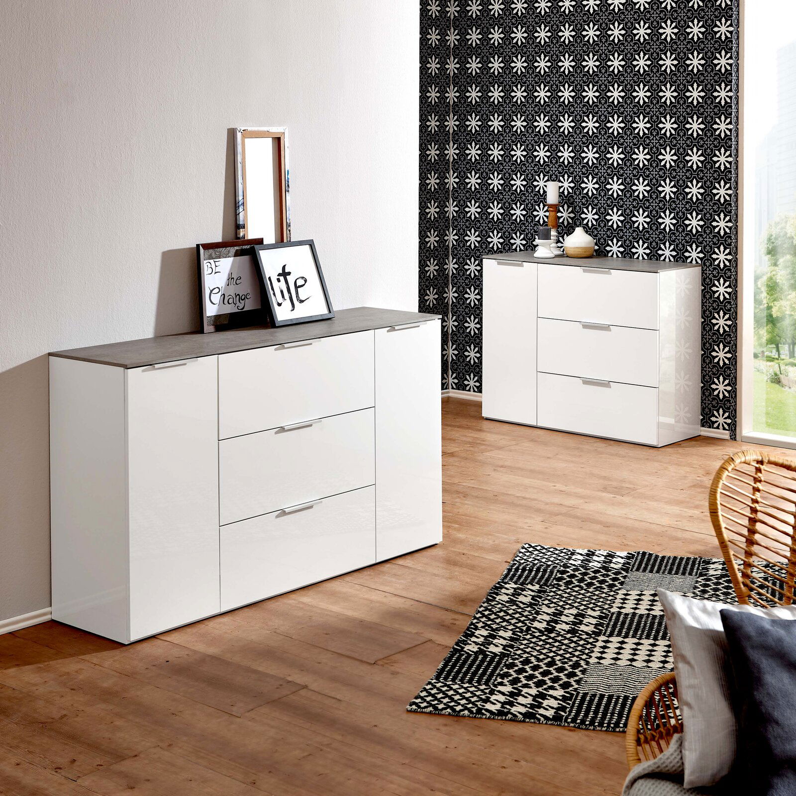 Modernes weißes Sideboard mit drei Schubladen und zwei Türen, in einem stilvoll eingerichteten Raum mit Holzboden und gemustertem Teppich, aus einer seitlichen Perspektive fotografiert.