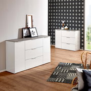 Modernes weißes Sideboard mit drei Schubladen und zwei Türen, in einem stilvoll eingerichteten Raum mit Holzboden und gemustertem Teppich, aus einer seitlichen Perspektive fotografiert.