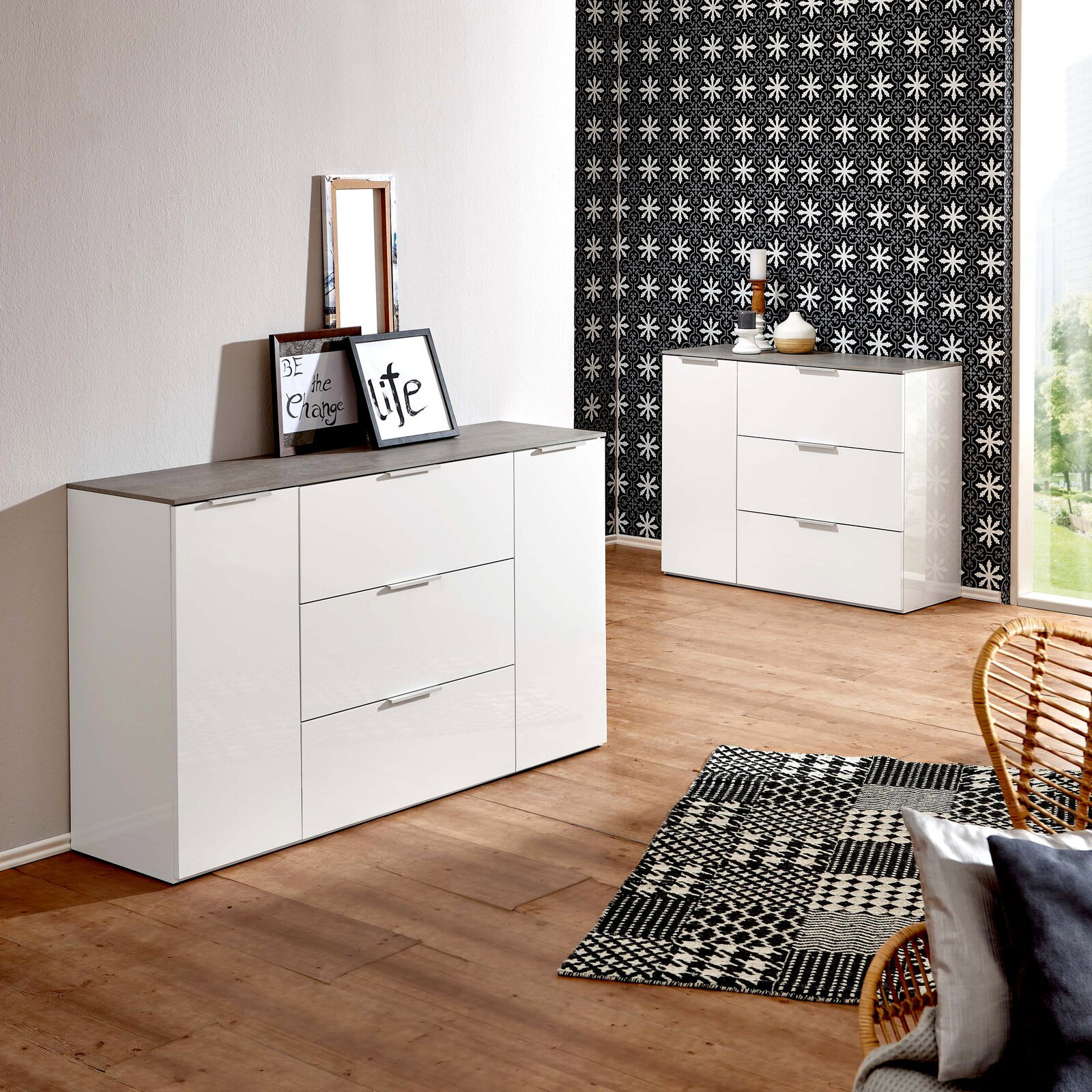 Modernes weißes Sideboard mit drei Schubladen und zwei Türen, in einem stilvoll eingerichteten Raum mit Holzboden und gemustertem Teppich, aus einer seitlichen Perspektive fotografiert.