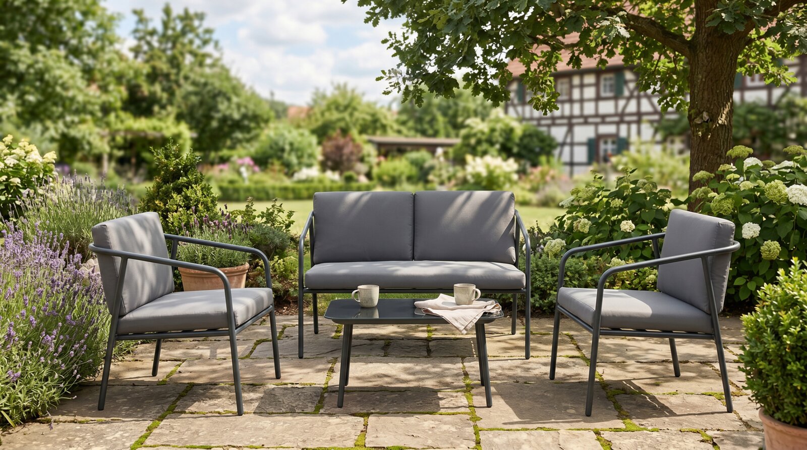 Garten-Lounge-Set mit dunkelgrauem Metallgestell und grauen Polstern: Zweisitzer-Sofa, zwei Sessel und niedriger Couchtisch auf Stein-Terrasse im grünen Garten, frontal aus Augenhöhe aufgenommen