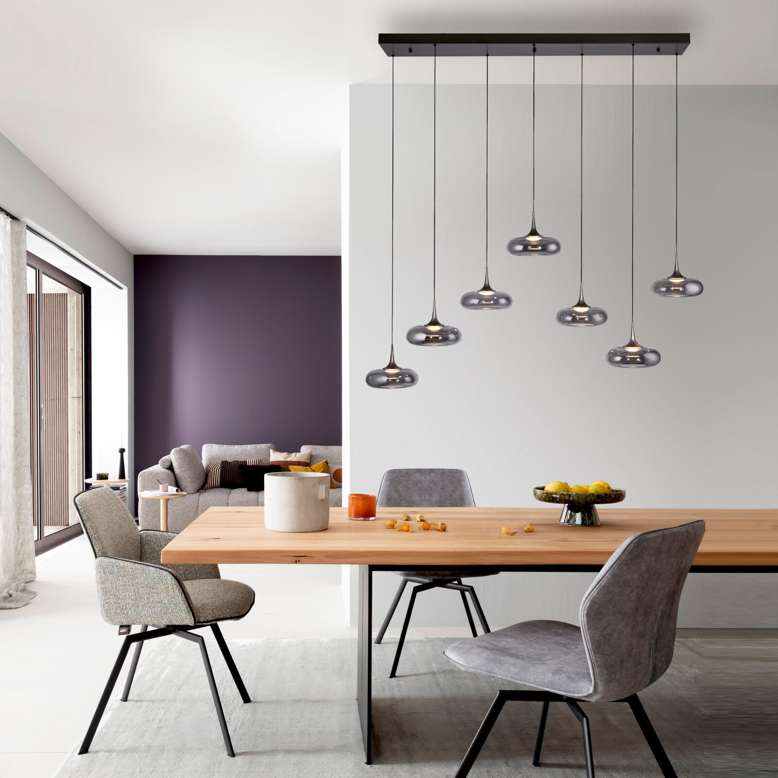 Moderne Mehrfach-Pendelleuchte mit sieben rauchgrauen Glas-Schirmen an schwarzer Deckenleiste über einem Holzesstisch im Wohnbereich, Frontalansicht in leicht erhöhter Perspektive