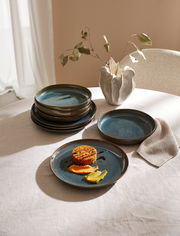 Ein Tafelset mit acht Teilen namens Breeze, bestehend aus eleganten, blauen Tellern auf einem Tisch. Die Perspektive zeigt die Teller von oben, mit einem dekorativen Gericht auf einem der Teller und einer stilvollen Vase im Hintergrund.