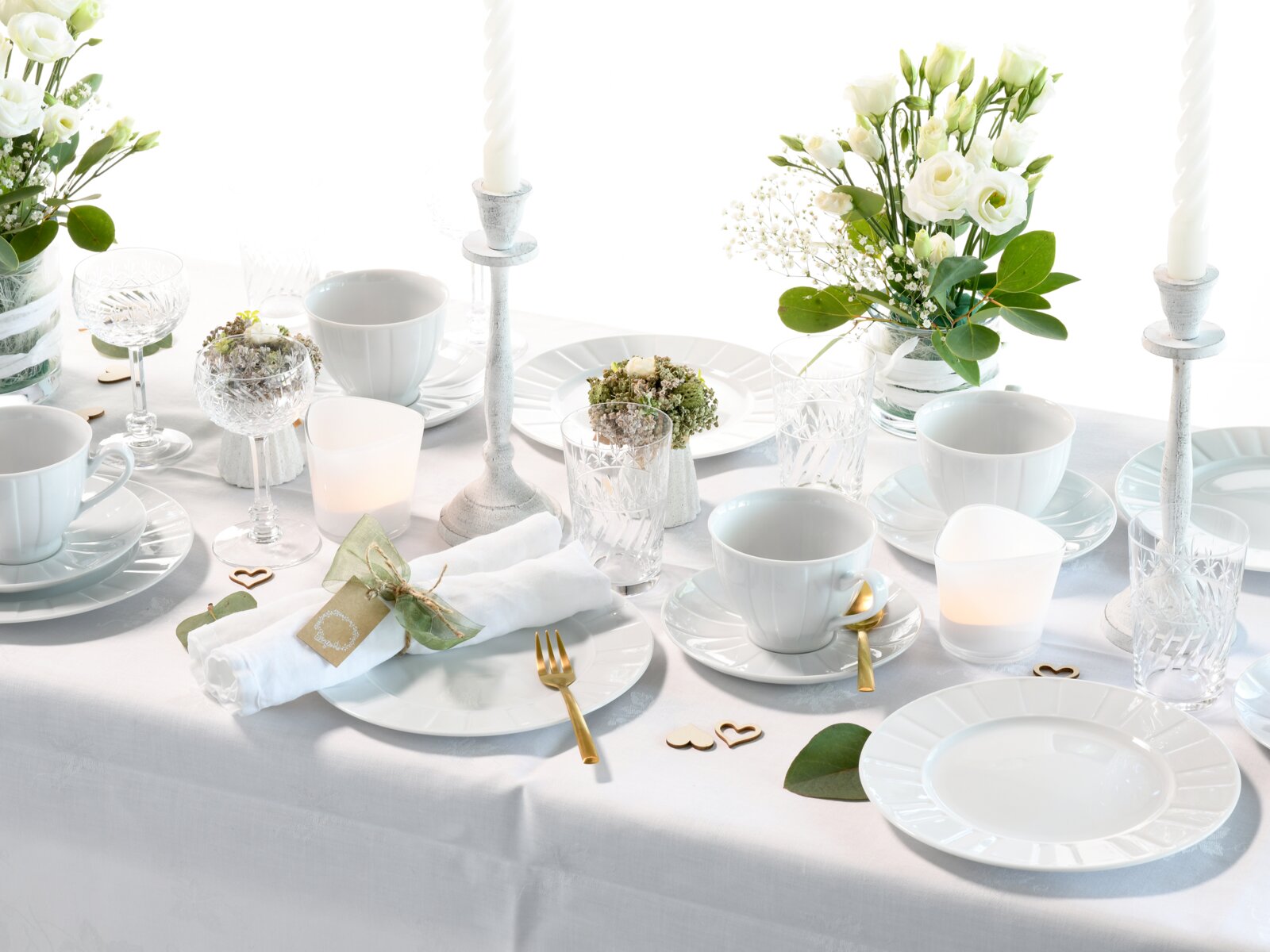 Festlich gedeckter Tisch in Weiß mit Porzellantellern und -tassen, Kristallgläsern, goldfarbenem Besteck, weißen Kerzen und Blumenarrangements, fotografiert aus leicht erhöhter schräger Perspektive (3/4-Ansicht).