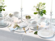 Festlich gedeckter Tisch in Weiß mit Porzellantellern und -tassen, Kristallgläsern, goldfarbenem Besteck, weißen Kerzen und Blumenarrangements, fotografiert aus leicht erhöhter schräger Perspektive (3/4-Ansicht).