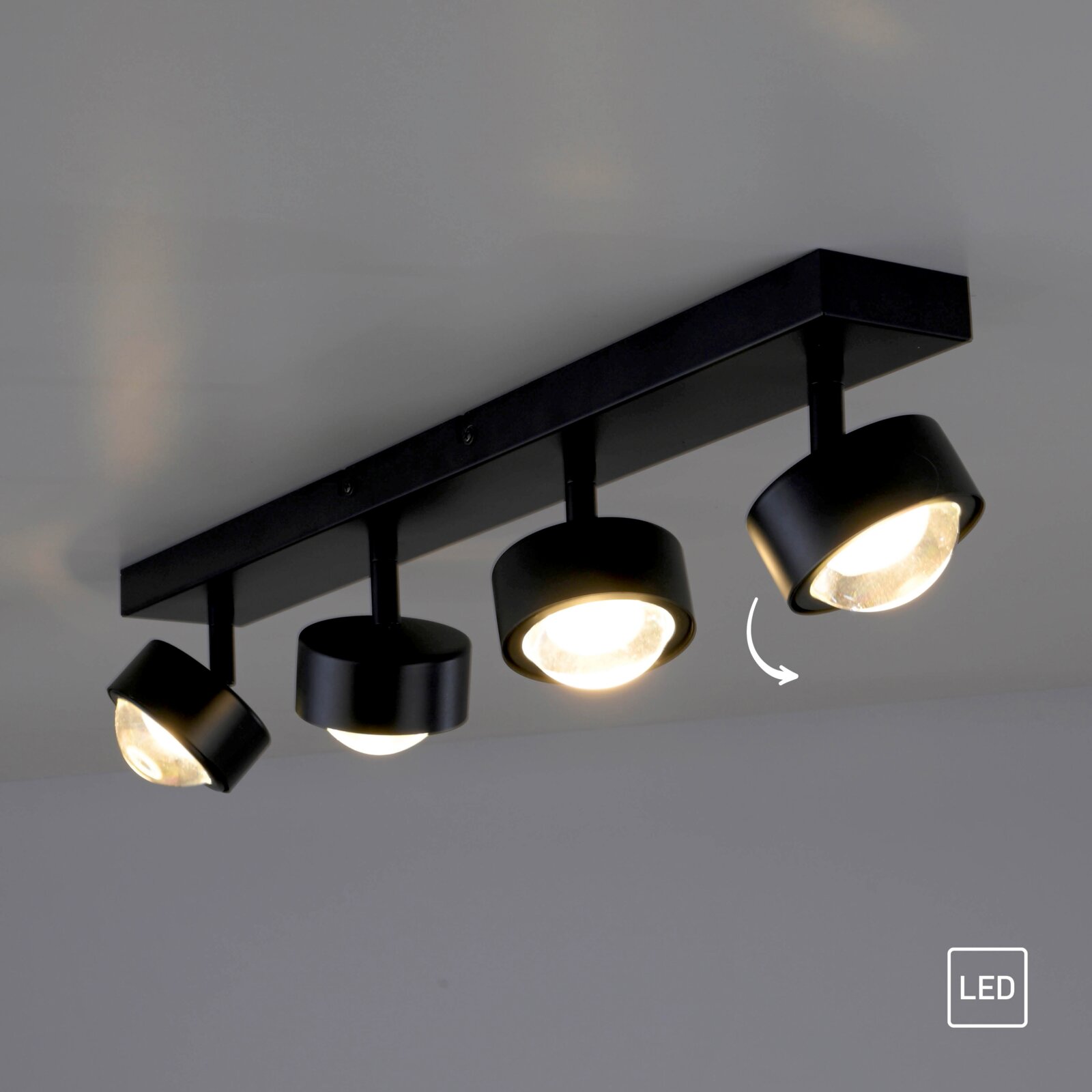 Schwarze, moderne LED-Deckenleuchte mit mehreren schwenkbaren Spotköpfen auf rechteckiger Schiene, warmweiß leuchtend, fotografiert aus leicht schräger Untersicht.