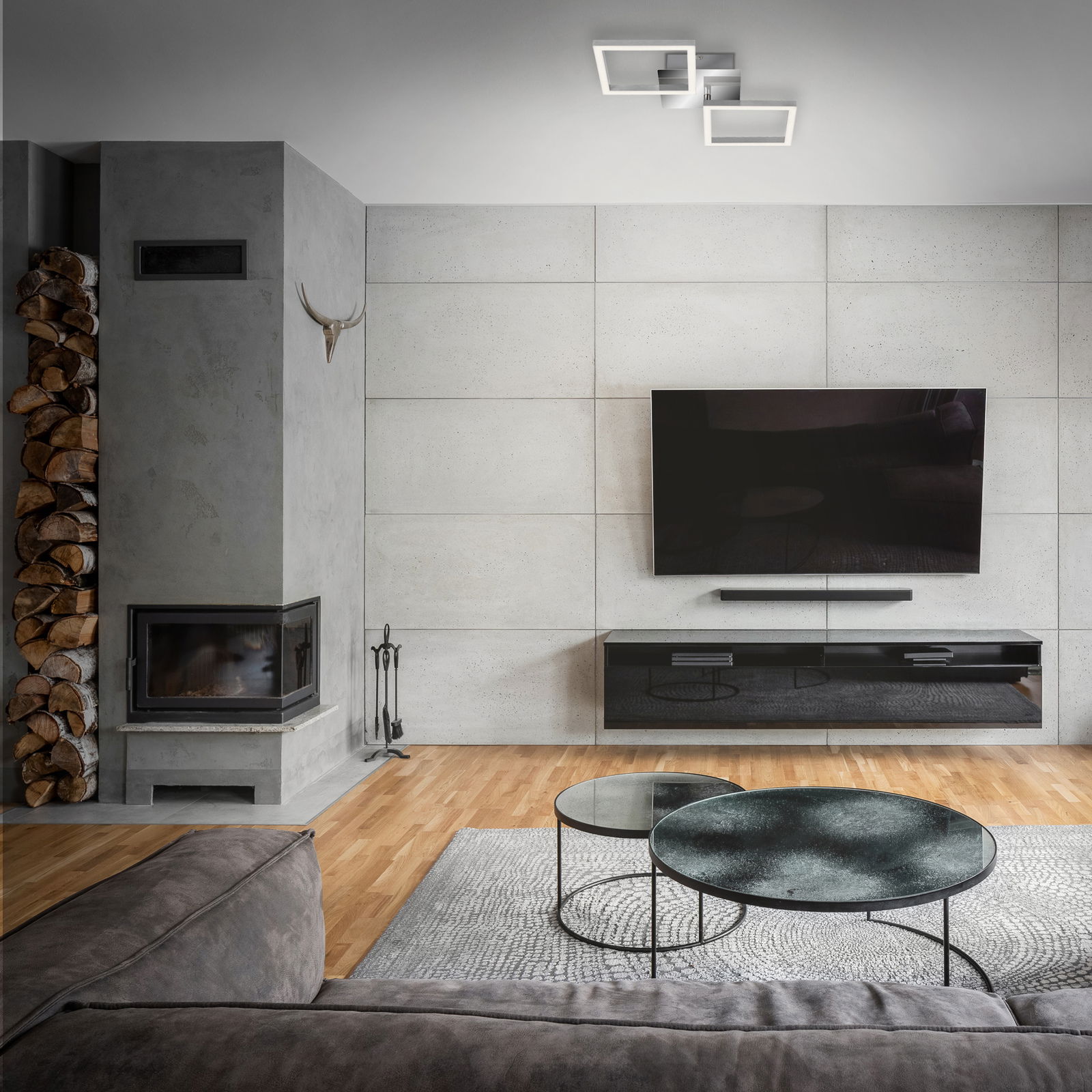 Moderne LED-Deckenleuchte in Chrom/Silber, von unten betrachtet in einem stilvollen Wohnzimmer mit grauer Wand und Kamin.